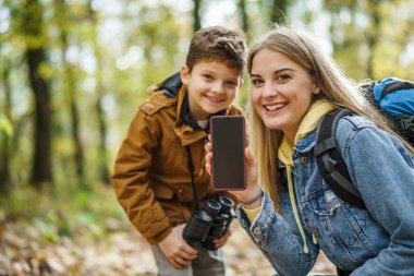 Happy mother and son are hiking in forest. They are using maps on smart phone. There is copy space on phone screen.