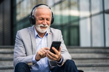 Mutlu kıdemli işadamı şirket binasının önünde işten sonra dinleniyor. Akıllı telefondan mesajlaşıyor..