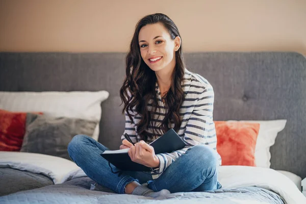 Young woman is sitting on bed at home and writing diary.