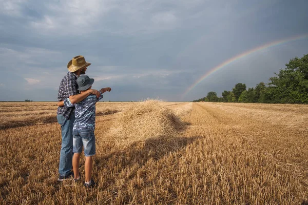 収穫が成功した後 父と息子は小麦畑に立っています 彼らは空に虹を見ている — ストック写真