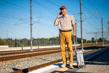 Happy senior man is standing at railway station and talking on smartphone. Senior people traveling.
