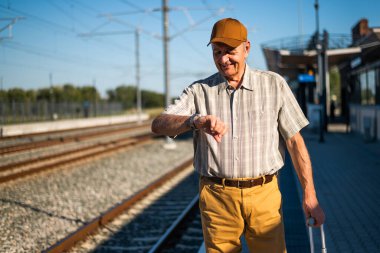 Mutlu kıdemli adam güneşli bir günde tren istasyonunda duruyor ve trenin gelişini bekliyor. O, kol saatine bakıyor. Son sınıflar seyahat ediyor..