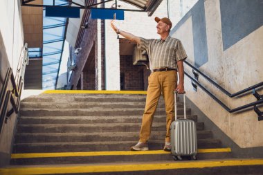 Mutlu kıdemli adam tren istasyonunda duruyor ve akıllı telefonuyla selfie çekiyor. Son sınıflar seyahat ediyor..
