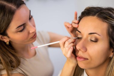 Güzellik uzmanı küçük bir fırça kullanarak başka bir kadına dikkatlice göz makyajı yapıyor. Makyaj yapan kadının gözleri kapalıyken sanatçı göz kalemi sürmeye odaklanıyor..