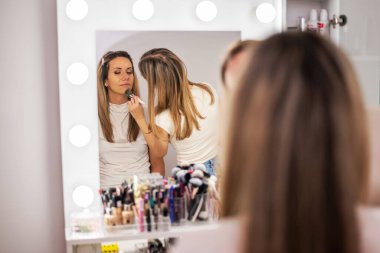 Güzellik uzmanı küçük bir fırça kullanarak müşteriye yüz pudrası uyguluyor. Yumuşak ışıklar ve ılık tonlar, bir makyaj sanatçısının iş yerindeki hassasiyetini ve kibarlığını vurgular..