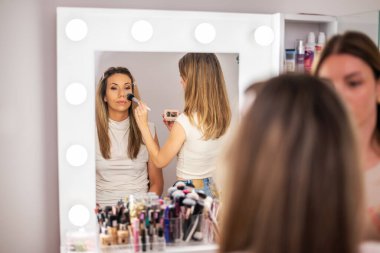 Bir makyaj sanatçısı güzellik salonundaki müşterisine yüz pudrası uyguluyor. Ressam ince bir fırça kullanıyor ve müşterisi sakince otururken işine odaklanmış görünüyor..