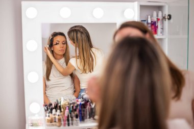 Güzellik uzmanı müşterinin yüzü üzerinde dikkatle çalışıyor. Sakince oturup makyaj seansının tadını çıkarırken müşterinin göz kirpiklerine rimel sürüyor..