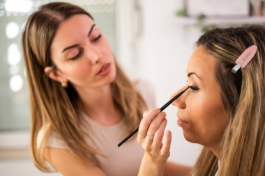 Güzellik uzmanı müşterinin yüzü üzerinde dikkatle çalışıyor. Sakince oturup makyaj seansının tadını çıkarırken müşterinin göz kirpiklerine rimel sürüyor..