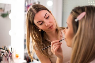İşteki güzellik uzmanı. Genç kadın güzellik stüdyosunda makyöz olarak çalışıyor. Bir müşterisine ruj sürüyor..