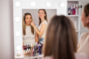 Mutlu makyaj sanatçısı tatmin olmuş müşterisiyle birlikte makyaj aynasının önünde duruyor. İkisi de gülümsüyor ve makyaj seanslarının keyfini çıkarıyorlar..