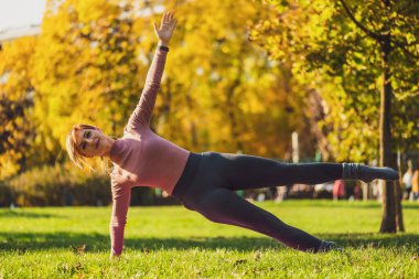 Sonbaharda parkta pilates yapan yetişkin bir kadın. Sağlıklı bir yaşam tarzı. Yan tahtada bacak tekmesi egzersizi var..