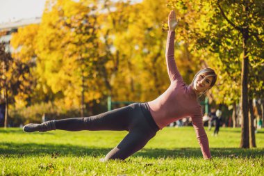 Sonbaharda parkta pilates yapan yetişkin bir kadın. Sağlıklı bir yaşam tarzı. Yan tahtada bacak tekmesi egzersizi var..