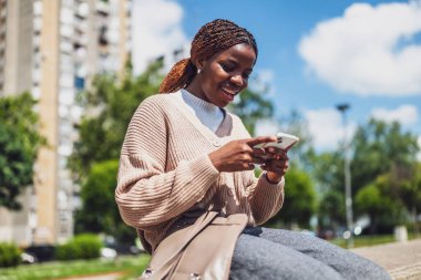 Telefonuyla mesajlaşırken gülümseyen Afro-Amerikan kadın, gün ışığında güneşleniyor, boş zamanları ve iletişimi sembolize ediyor.