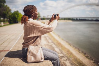 Genç, neşeli siyah kadın akıllı telefonuyla fotoğraf çekiyor. Şehirdeki nehrin kenarında oturuyor..