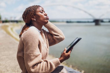 Genç endişeli siyah kadın dijital tablet kullanıyor. Şehirdeki nehrin kenarında oturuyor..