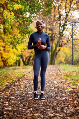 Atletik giyinmiş bir kadın, elinde bir su şişesi tutarken gülümser, yapraklarla kaplı bir patikada durur, parlak sonbahar yapraklarıyla çevrilidir..