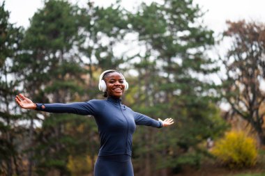 Rahat ve mutlu görünen siyah bir kadın, kollarını açmış parkta duruyor. Antrenmana hazır..