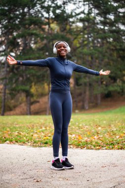 Rahat ve mutlu görünen siyah bir kadın, kollarını açmış parkta duruyor. Antrenmana hazır..