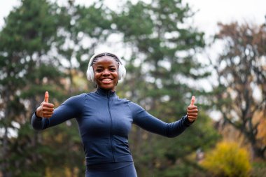 Koşuya hazır siyah bir kadın kışın parkta durur, rahat ve mutlu görünür, başparmaklarını kaldırır..
