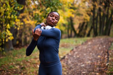 Genç siyah kadın sonbaharda parkta egzersiz yapıyor. Koşmadan önce vücudunu esnetiyor..