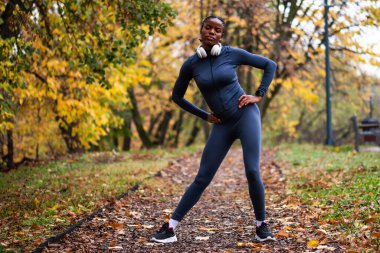 Genç siyah kadın sonbaharda parkta egzersiz yapıyor. Koşmadan önce vücudunu esnetiyor..