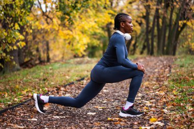 Genç siyah kadın sonbaharda parkta egzersiz yapıyor. Koşmadan önce vücudunu esnetiyor..