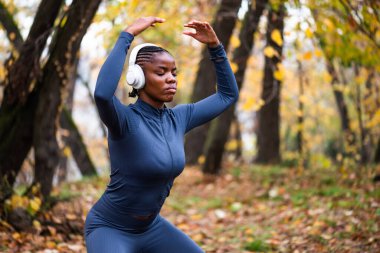 Genç siyah kadın sonbaharda parkta Tai Chi egzersizi yapıyor. Kablosuz kulaklık takıyor..