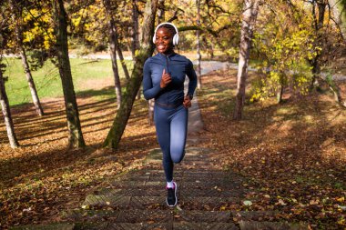 Canlı sonbahar renkleriyle dolu bir orman parkında spor kıyafetleriyle gülümseyen bir koşucu müzik dinlerken açık havada merdivenlere tırmanıyor..