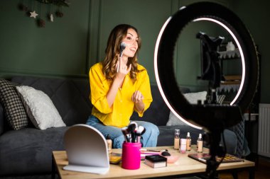 Bir kadın, bir kanepede oturup makyaj yaparken güzellik dersi alıyor. Yüzük ışığıyla aydınlatılıyor ve bir masanın üzerinde kozmetik ürünlerle çevrili..