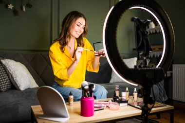 Bir kadın, bir kanepede oturup makyaj yaparken güzellik dersi alıyor. Yüzük ışığıyla aydınlatılıyor ve bir masanın üzerinde kozmetik ürünlerle çevrili..