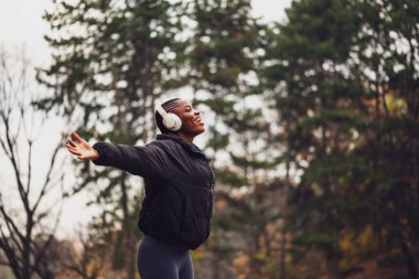 Rahat ve mutlu görünen siyah bir kadın, kollarını açmış parkta duruyor. Kulaklıkla müzik dinliyor ve soğuk havada doğanın tadını çıkarıyor..