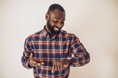 Rahat gömlekli neşeli siyah adam duvarın önünde duruyor ve kol saatine bakıyor..