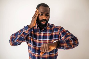 Rahat gömlekli endişeli siyah adam duvarın önünde duruyor ve kol saatine bakıyor. Geç kalacak..