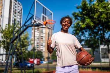 Afrika kökenli Amerikalı bayan oyuncu bir top tutuyor ve yaz antrenmanı sırasında başparmağını kaldırıyor. Aktif yaşam tarzı ve şehir sporları.