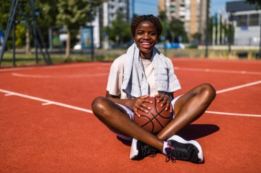 Genç Afrikalı Amerikalı kadın güneşli şehir basketbol sahasında oturuyor, neşeli bir gülümsemeyle bir top tutuyor, neşeyi ve aktif yaşam tarzını temsil ediyor..