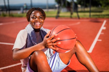 Genç siyah kadın basketbol sahasında oturuyor. Topu elinde tutuyor..