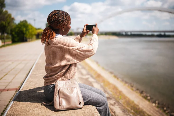 Genç, neşeli siyahi kadın nehir kenarında oturuyor ve fotoğraf çekiyor..