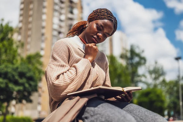 Afro-Amerikalı kadın şehir parkında dinleniyor, derin düşüncelere dalmış ve günlüğünü yazıyor..