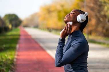 Sporcu olan siyahi bir kadın, güneşli bir günde parkta koşu pistinde durarak, koşudan sonra terleyerek yüzünü siliyor..
