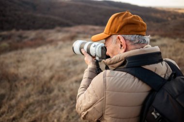 Şapka, yastıklı ceket ve sırt çantası takmış bir adam dağda dikiliyor, fotoğraf makinesi merceğiyle fotoğraf çekiyor..