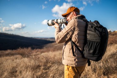 Şapka, yastıklı ceket ve sırt çantası takan bir adam dağda dikiliyor, kamera merceğiyle manzarayı veya vahşi yaşamı fotoğraflıyor..