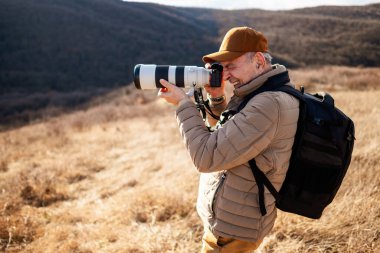Şapka, yastıklı ceket ve sırt çantası takan bir adam dağda dikiliyor, kamera merceğiyle manzarayı veya vahşi yaşamı fotoğraflıyor..