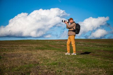 Kahverengi şapkalı, yastıklı ceketli ve sırt çantalı bir adam vahşi yaşam ya da manzara fotoğrafları çekiyor. Etrafı yuvarlanan tarlalar ve dramatik beyaz bulutlarla çevrili..