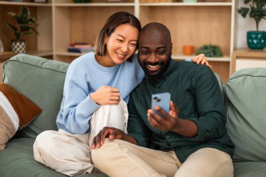 Mutlu ve sevgi dolu çok ırklı çift evde dinlenirken neşeli bir anı paylaşıyor. Akıllı telefon kullanıyorlar..