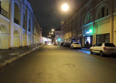 Gece caddelerinin manzarası, sarı ışıkla aydınlatılmış, İmparatorluk tarzı sarı binalar, arka plandaki arabalar.          