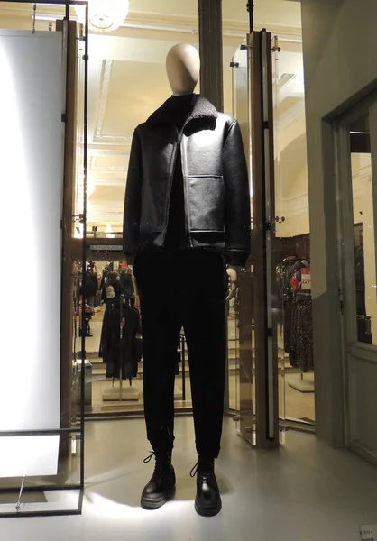 Male mannequin with a white head dressed in a black sheepskin coat, black trousers and boots in the window of a clothing store