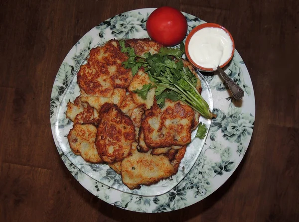 a dish full of freshly baked potato pancakes on a transparent dish, on a round white-green napkin, next to a bowl with sour cream and a tomato