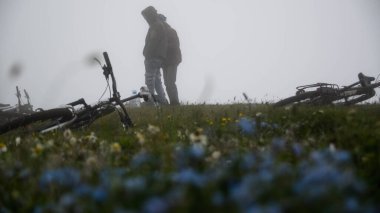 Odaklanamayan iki adamın siluetleri sislerin arasında duruyor. Bisikletlerin ve çiçeklerin arkasında.