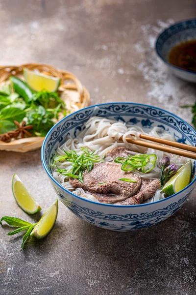 Vue rapprochée de pho bo dans un bol traditionnel, garni de basilic, menthe, citron vert, sur fond de béton — Photo