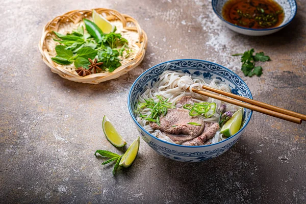 Soupe vietnamienne traditionnelle aux nouilles pho dans un bol, garnie de basilic, menthe, citron vert, sur fond de béton — Photo